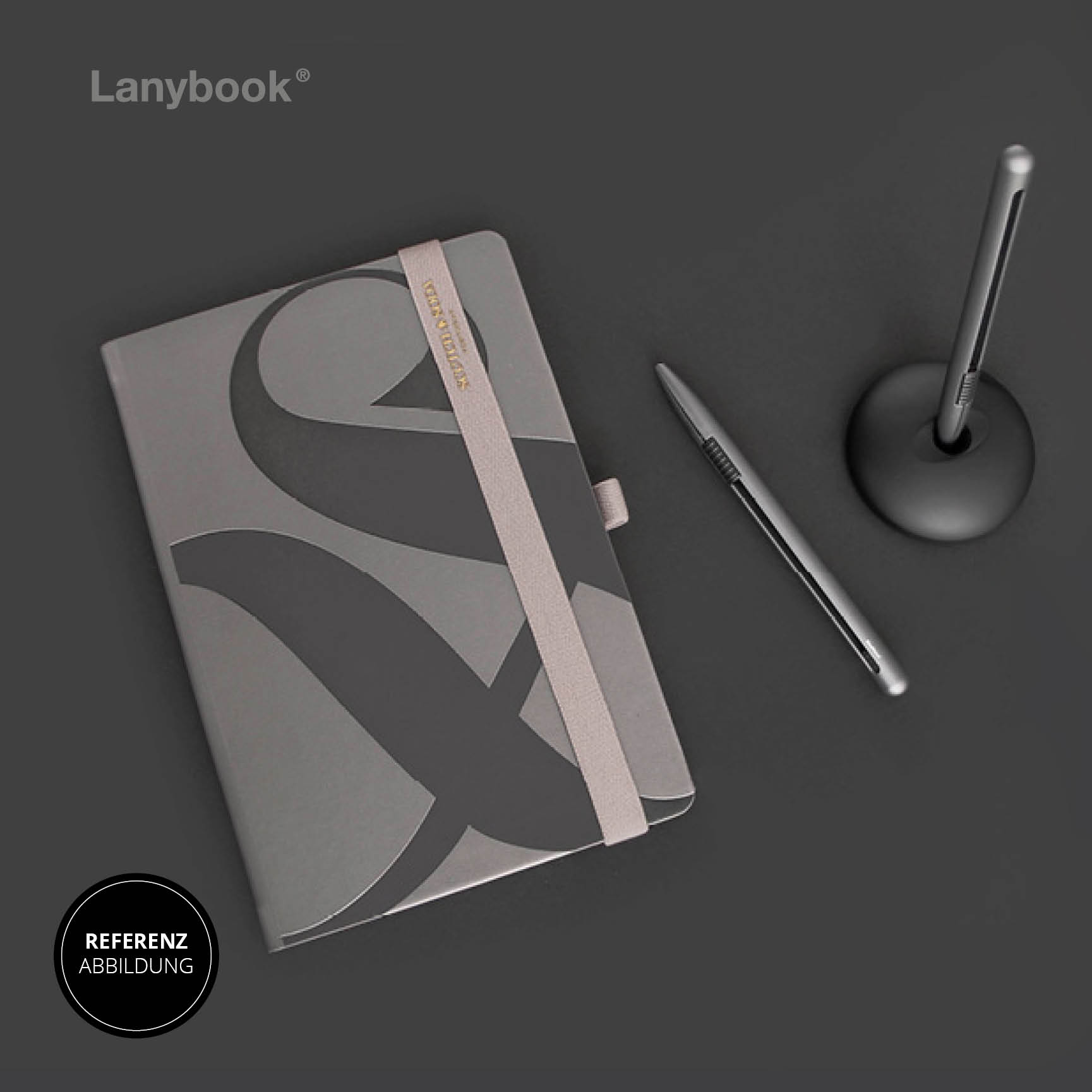 Graues Notizbuch mit schwarzem Design, beigem Lanyband und Stiftschlaufe, daneben zwei Stifte auf dunklem Hintergrund.