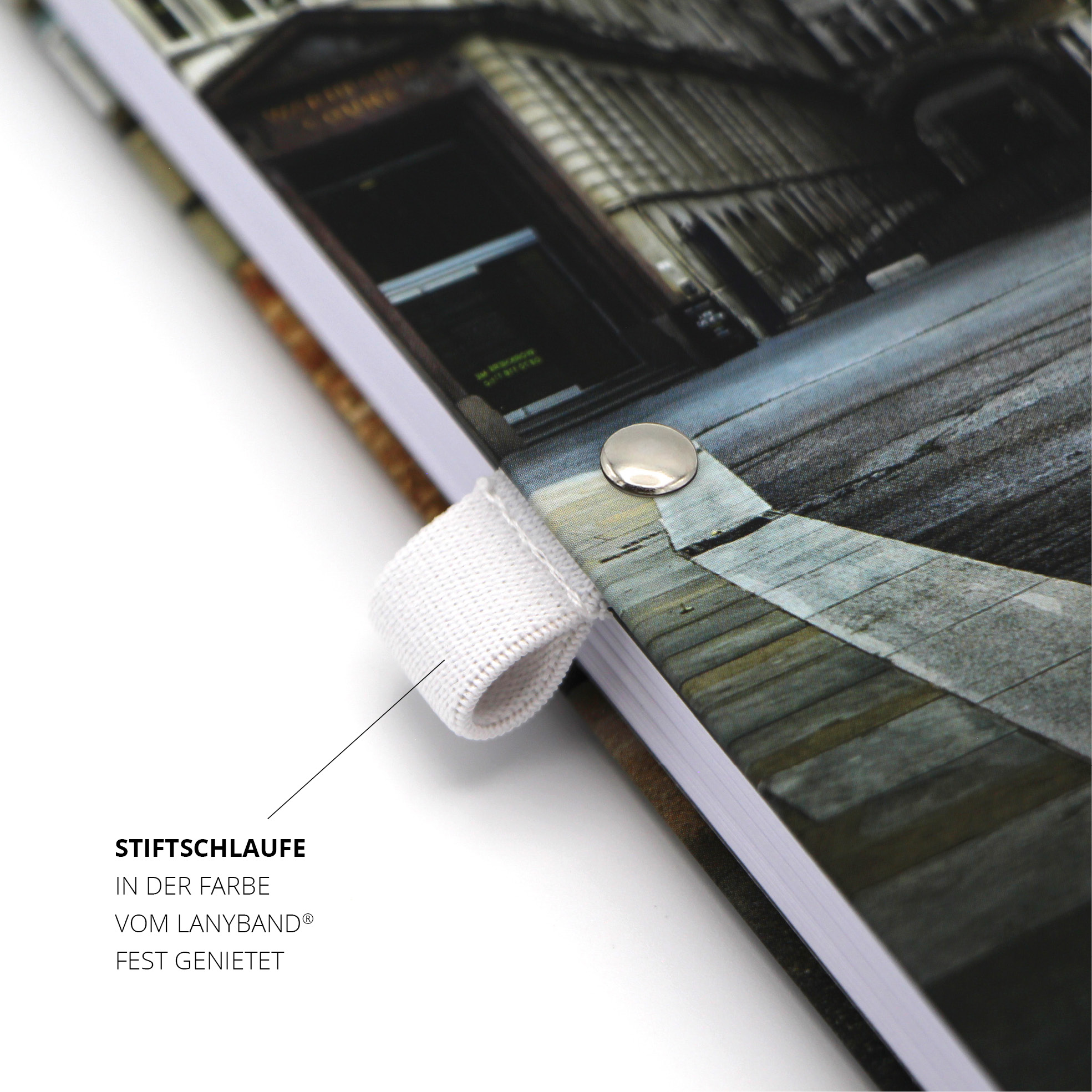 Weiße Stiftschlaufe am Notizbuchrücken, silbern vernietet, urbanes Motiv im Hintergrund.