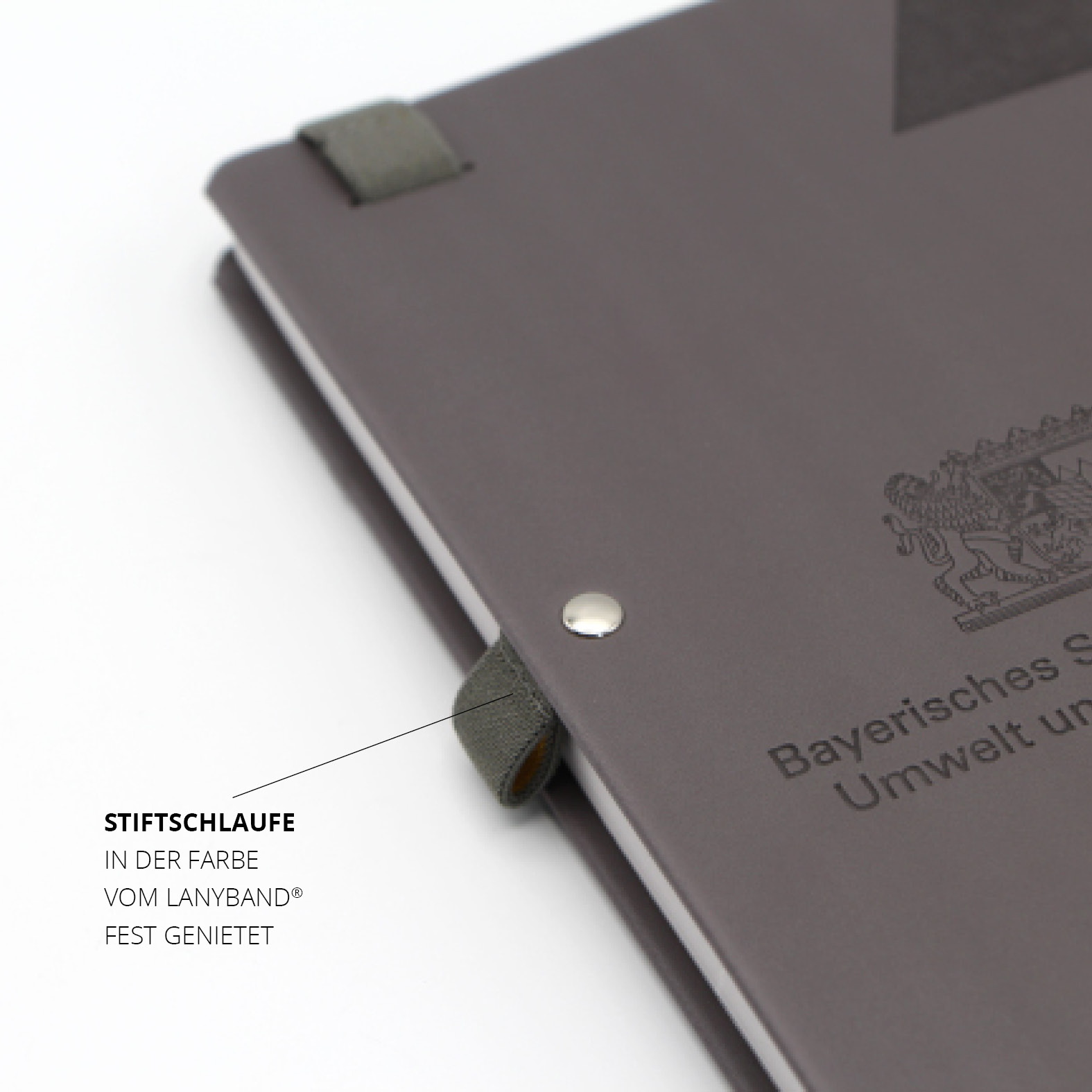 Detailansicht der farblich abgestimmten Stiftschlaufe, fest vernietet am Notizbuchcover mit geprägtem Logo des Bayerischen Staatsministeriums.